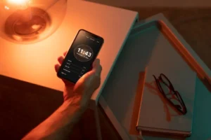 شخص يمسك هاتفه بجانب دفتر ونظارة تحت إضاءة دافئة