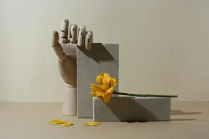 يد خشبية بجانب زهرة صفراء فوق مجسمات minimal في مشهد هادئ يعبر عن الثقل النفسي